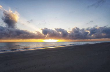 Görüntü, okyanusun üzerinde nefes kesici bir gün doğumunu dramatik bulutların kumlu kumsalda uzun gölgeler oluşturduğunu gösteriyor.. 