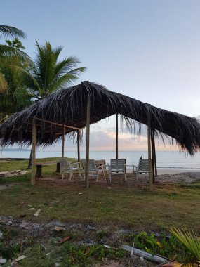 Görüntü, kumlu bir sahilde birkaç sandalyesi olan sazdan çatılı bir kulübeyi gösteriyor..