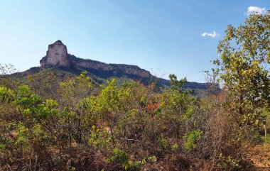 Sahne, gökyüzüne karşı keskin bir şekilde yükselen belirgin bir kaya oluşumunun yer aldığı engebeli bir manzarayı tasvir ediyor..