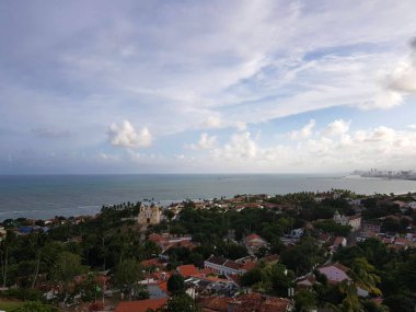 Görüntü bir sahil şehrinin panoramik manzarasını gösteriyor. Ön planda, birçok palmiye ağacına sahip yoğun tropikal bitki örtüsü vardır.. 