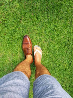 Sahne yeşil çimlerin üzerinde duran bir çift bacağı gösteriyor. Bir ayağı kahverengi bir ayakkabı giyer, diğer ayağı beyaz bir terlik giyer..