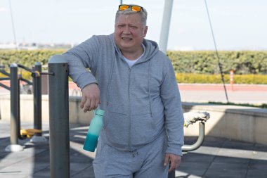 Yaşlı bir adam dışarıda bir fitness parkında mola veriyor, sağ elinde turkuaz bir su şişesi tutuyor. Arka planda açık spor malzemeleri var.