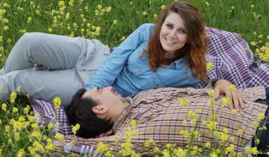 Genç bir çift ekose bir battaniyenin üzerinde çiçek açan bir çayırda dinleniyor. Erkek onun yanında dinlenirken kadın kameraya gülümsüyor. Romantik ve neşeli bir bahar anı doğada, aşk teması için ideal.                               
