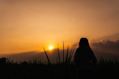 Asyalı bir kadın dramatik bulutlarla gün batımını seyrederken sahada duruyor. arkaplan görünümü kavramı.