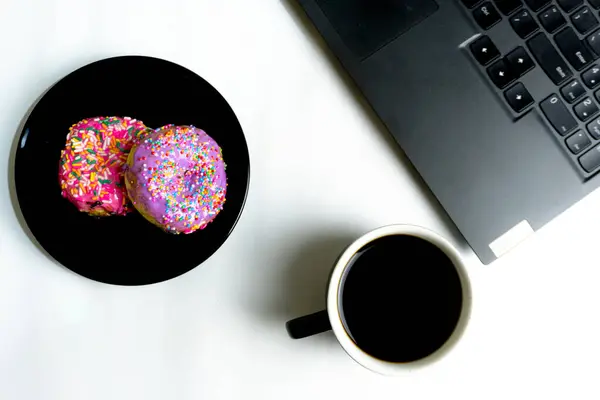 Görüntü bir fincan kahve, iki şekerli çörek ve bir dizüstü bilgisayar gösteriyor. Çörekler siyah tabakta. Görüntü yukarıdan aşağıya doğru alınır ve arkaplan beyazdır.