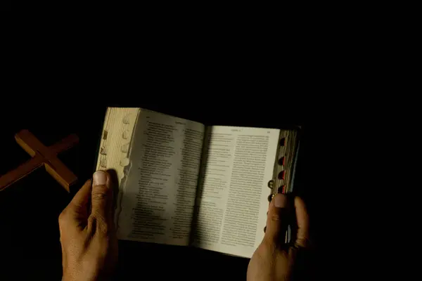 Karanlık arka planda inanç ve maneviyatı simgeleyen açık bir İncil ve ahşap bir haç tutan bir kişiyi tasvir ediyor.