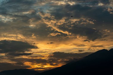 Bu görüntü bir dağ sırasının üzerinde güzel bir gün batımı yakalar. Gökyüzü bulutlu gökyüzü ile canlı turuncu ve sarıdır. Güneş dağların ardında batıyor, manzaranın üzerine uzun gölgeler bırakıyor..