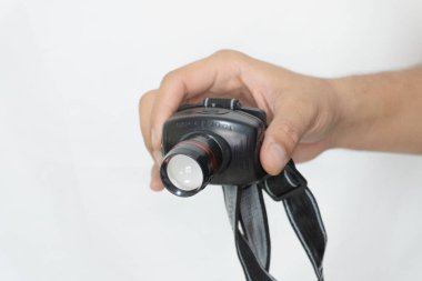 a person's hand holding a black headlamp. The headlamp has a red stripe and a white lens. The hand is holding the headlamp in front of a white background. clipart