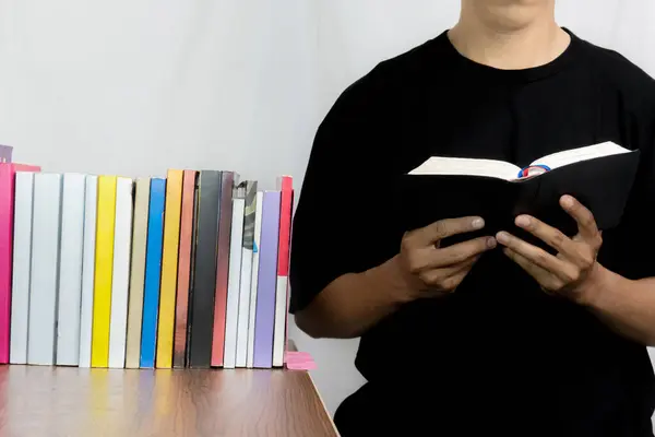 Biri pembe bir kitabı açık tutuyor ve arka planda tahta bir masanın üzerinde bir sıra kitap var. Arkadaki kitaplar çok renklidir. Siyah bir gömlek giyiyor..