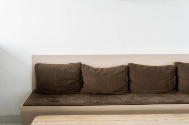 Düz beyaz bir duvarın önünde kahverengi bir kanepe oturuyor. Kanepede dört tane kahverengi yastık var.