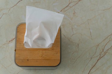 Bambu kapaklı kare doku kutusu açık bej mermer bir yüzeye oturur. Kutudan tek bir mendil çıkarıldı..