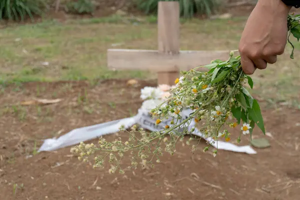 Bir insanın eli küçük bir buket kır çiçeğini mezarın önünde basit bir ahşap haçla tutar.