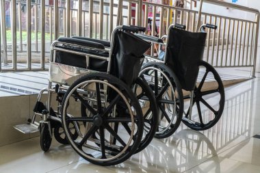 İki siyah tekerlekli sandalye, parlak fayanslı bir zeminde metal korkuluklara karşı yan yana durur..