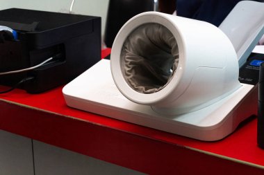 Siyah yazıcı ve güç kablolarının yanına kırmızı bir yüzeye yerleştirilmiş beyaz otomatik bir tansiyon monitörü..