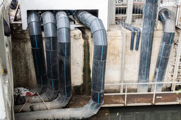 Mavi çizgili çoklu siyah borular, küçük bir kanal üzerinde, pürüzlü bir şehir ortamında dikey ve yatay olarak ilerler..