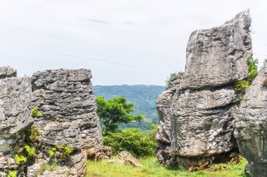 İki büyük kayalık sütun, parlak ve bulutlu bir gökyüzünün altındaki yeşil dağların manzarasını oluşturur..