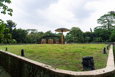 Ağaçlarla dolu yeşil bir parkın ortasında, bir barınak ve şemsiyesi olan boş bir çitli alan..