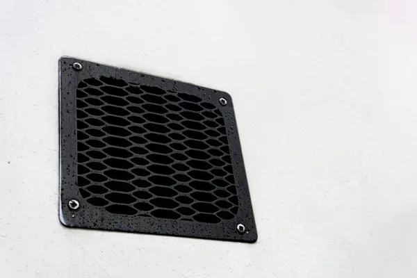 A black metal grate or vent cover with an oval pattern, mounted on a light-colored, possibly white, surface.