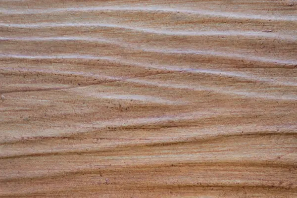 A close-up of a textured, light brown wooden or wood-like surface with horizontal, wavy patterns.