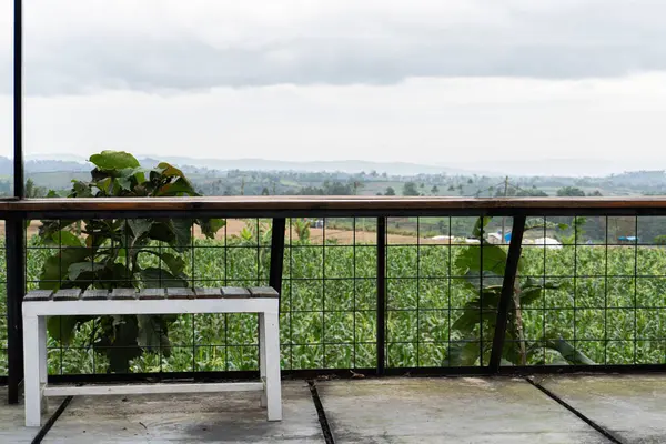 Yükseltilmiş bir platformdan sakin bir manzara yeşil bir tarlaya bakan beyaz bir bank ve bulutlu bir gökyüzünün altındaki uzak tepeler..