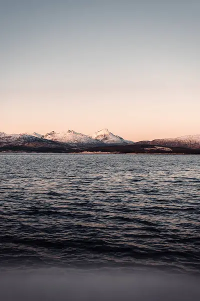 Tromso, Norveç 'teki fiyordun yanındaki karlı arktik dağlarının üzerinde gün batımı.