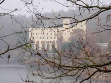 Orlik Şatosu sisli bir sonbahar gününde aynı isimde bir barajın üzerinde yükseliyor. Yürüyüşçüler için popüler bir yer, dallar arasında alışılmadık bir manzara.