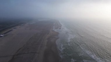 Dalgaların ve kumlu bir kıyı şeridinin yer aldığı sakin, sisli bir sahil manzarası sükuneti ve doğanın güzelliğini çağrıştırıyor. Kuzey Denizi, Kış, Hollanda
