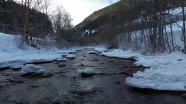 Sakin bir kış sahnesi, akan bir nehir, karla kaplı kıyılar ve arka planda görkemli dağlar.