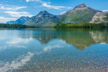 Rocky Dağları Kanada 'daki Waterton Lakes Ulusal Parkı' nın aşağısındaki göle yansımaktadır. Gökyüzü, beyaz bulutları olan muhteşem bir mavi. Arka planda Galler Prensi Oteli var ve gölün sığ kenarında renkli kayalar var.. 