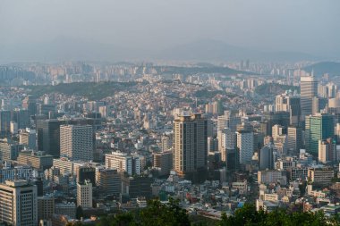 Seul gökdelenlerinin ve geleneksel binaların sisli bir gökyüzü altında şehir yaşamını aydınlatan dinamik bir karışımını gözler önüne seriyor.