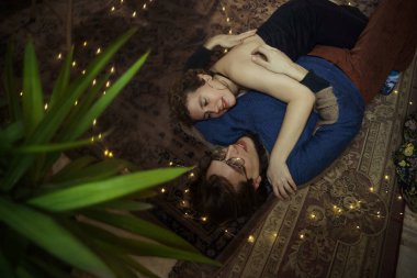 İki insan sıcak bir atmosferde dekoratif ışıklarla çevrili yumuşak bir halının üzerinde uzanırken samimi bir anı paylaşır..