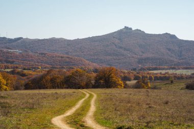 Dolambaçlı bir toprak yol, arka planda tepeler ve yukarıda açık mavi bir gökyüzü olan renkli bir sonbahar manzarasına çıkar..