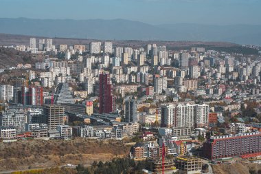 Şehir siluetinde açık mavi gökyüzü altında modern binalar ve devam eden inşaat projelerinin bir karışımı yer alıyor..