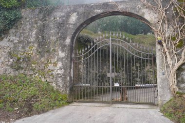 Bu anımsatıcı görüntü, yıpranmış taş bir duvarın içinde kısmen sisle kaplanmış dövülmüş demir bir kapı içeriyor. Dekoratif dikenlerle süslenmiş kapı kapalı duruyor ve ardında saklı bir dünya olduğunu gösteriyor. Sadık bir köpek içeride dinlenirken görülebilir.