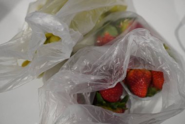 Bu canlı görüntü, temiz plastik poşetlere doldurulmuş taze meyve çeşitlerini gösteriyor. Odak noktası dolgun çilekler. Parlak kırmızı renkleri çevredeki yeşilliklerle çok uyumsuz. Arka planda, yeşil üzüm izleri var.