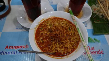 Danau Toba, 09 Kasım 2024. Kareli masa örtüsünde koyu renkli bir içecek içeren bir kase baharatlı hazır erişte servis ediliyor. Sumatra Utara, Endonezya.