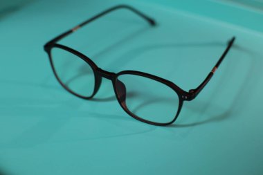 Bir çift siyah çerçeveli gözlük açık mavi bir yüzeye dayanır. Gözlüklerin kare şeklinde ve parlak bir malzemeden yapılmış. Gözlüklerin yansıması yüzeyde görülebilir..