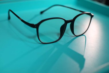 A pair of black-framed glasses rests on a light blue surface. The glasses have a square shape and are made of a shiny material. The reflection of the glasses can be seen on the surface. clipart