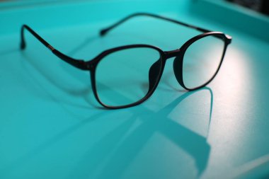 A pair of black-framed glasses rests on a light blue surface. The glasses have a square shape and are made of a shiny material. The reflection of the glasses can be seen on the surface. clipart