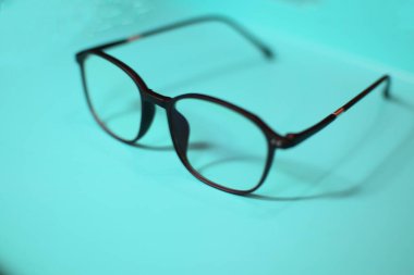 Bir çift siyah çerçeveli gözlük açık mavi bir yüzeye dayanır. Gözlüklerin kare şeklinde ve parlak bir malzemeden yapılmış. Gözlüklerin yansıması yüzeyde görülebilir..