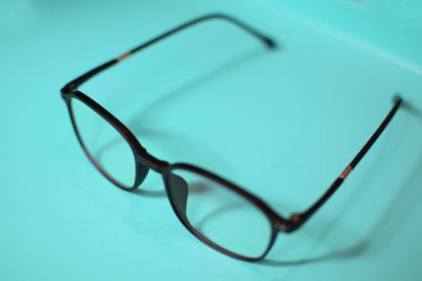 Bir çift siyah çerçeveli gözlük açık mavi bir yüzeye dayanır. Gözlüklerin kare şeklinde ve parlak bir malzemeden yapılmış. Gözlüklerin yansıması yüzeyde görülebilir..