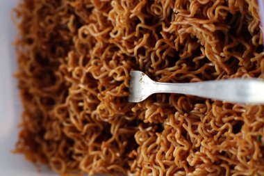 Beyaz bir kabın içinde pişmiş kahverengi erişteyi gösteren yakın plan bir fotoğraf. Eriştenin içine gümüş bir çatal yerleştiriliyor, yemeye hazır olduğunu gösteriyor. Eriştenin dokusu ve ayrıntıları vurgulanmış..