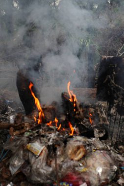 Yakın çekim, alevler ve yoğun dumanla havaya yükselen bir çöp yığınını gösteriyor. Atık madde plastik ve diğer döküntüleri içeriyor gibi görünüyor. Arka plan, kararmış ağaç gövdeleri ve çalılıkların yakınında bir orman ortamı gösteriyor.