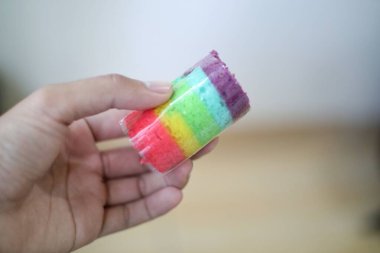 Bir el zarif bir şekilde canlı bir gökkuşağı katmanlı pasta tutar, her bir renk farklı ve titizlikle katmanlıdır. Minyatür kekin parlak renkleri görsel olarak çekici ve lezzetli bir ziyafet yaratır..