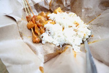 Yakın plan çekimde satay ve pirinçten arta kalanları buruşuk kahverengi kağıt ambalajında resmediliyor. Sahne basit ama sıradan bir yemek hissini uyandırıyor. Odak noktası yemeğin dokuları ve renkleri..