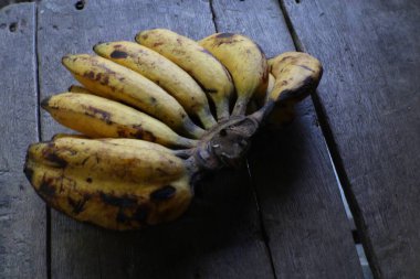Olgun muz kümesi yıpranmış ahşap bir yüzeyde bulunur, zengin sarı renklerini ve ince koyu beneklerini sergiler. Görüntü kırsal ve doğal bir hissi çağrıştırıyor..