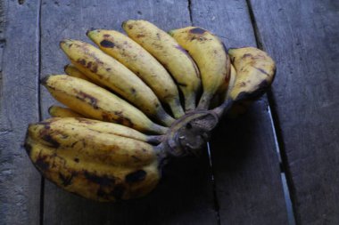Bir grup olgun muzun yakın plan görüntüsü sarı renklerini ve ahşap arka planlarını gözler önüne seriyor. Görüntü, doğal bir tazelik ve basit bir bolluk hissi uyandırıyor..