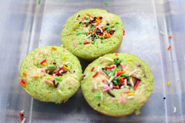 Üç pastel yeşil kek, gökkuşağı parçacıkları ve kremalı dolgu malzemeleriyle süslenmiş, berrak plastik bir kapta dinleniyorlar. Canlı ve neşeli bir görüntü..