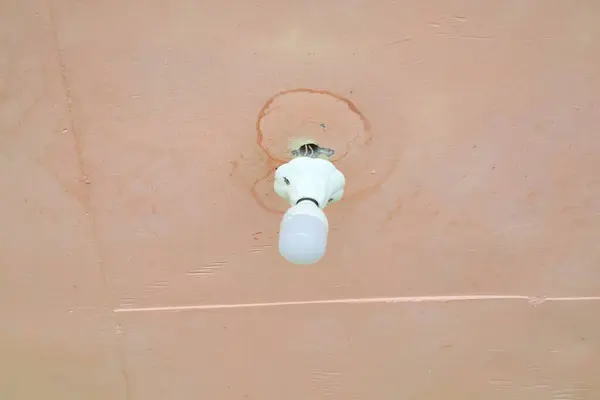 Boyalı bir yüzeye yakın çekim tavan lambası su hasarı ve yaşlanmayı gösteriyor. Ampul, yüzeydeki düzensizlikleri vurgulayan yumuşak bir parıltı yayar..