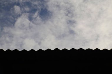 Yumuşak, dağınık bulutlarla dolu parlak bir gökyüzüne karşı bükülmüş metal bir çatının katıksız silueti. Çatının dalgalı deseni gökyüzünün havadar genişliğiyle çelişiyor ve görsel olarak ilginç bir kompozisyon oluşturuyor..